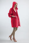 A person wearing a bright red, water-resistant **looselyboho VIVA MAGENTA WINDBREAKER** with the hood up, beige leggings, and beige high-heeled sandals stands against a plain white background. The individual has their hands in the coat pockets and is looking downward.