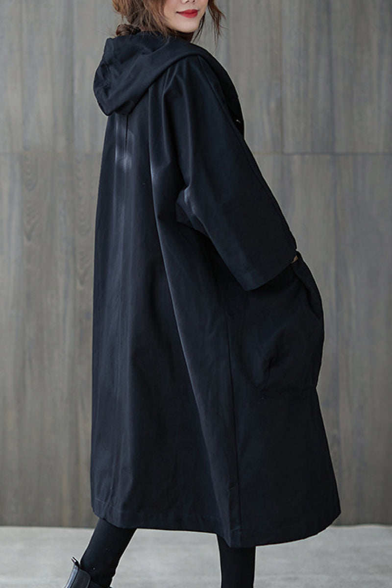 A person is standing sideways wearing the looselyboho Black One Size Fits All Oversized Waterproof Windbreaker Jacket with their hands in their pockets. The oversized fit of the jacket provides extra wind protection. The background consists of a plain, gray, wooden surface.