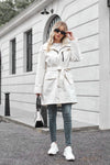 A blonde woman wearing sunglasses, a BELTED FROST WHITE WINDBREAKER by looselyboho, blue jeans, and white sneakers stands on a street in front of a white building with black windows. She is holding a black and white handbag and lightly touching her sunglasses.