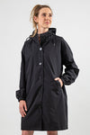 A person with long hair stands against a plain backdrop wearing a stylish rain-jacket featuring the looselyboho ONYX BLACK WINDBREAKER with an adjustable hood and elastic-cuffed sleeves. The person's hands are in their windbreaker's pockets, and their expression is neutral.