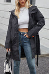 A woman with blonde hair wearing sunglasses is dressed in a stylish BELTED MIDNIGHT BLACK WINDBREAKER by looselyboho, a white crop top, and blue jeans. She is holding a black and white handbag and standing in front of a modern, light-colored building.