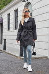 A woman with long blonde hair wears a looselyboho BELTED MIDNIGHT BLACK WINDBREAKER tied at the waist, light blue jeans, and white sneakers. She carries a small black handbag and sports dark sunglasses. She is walking down a grey street with a white building in the background.